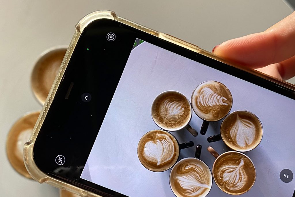 Close-up of a smartphone capturing a top-down photo of six cappuccinos with latte art arranged in a circle on a white table. The image represents visually sharing aesthetic content, imagery for both website and social media posts.