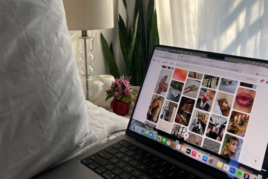 A laptop resting on a cozy bed displays a Pinterest feed filled with lifestyle and beauty images, set against soft natural light and a minimal bedroom backdrop. The scene reflects how social media platforms are often browsed casually and visually, contrasting with the more intentional, content-rich experience of a well-built website.