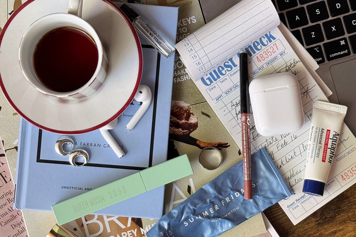 Flat lay of a cluttered workspace featuring a half-full cup of tea, AirPods, rings, lip balm, beauty products, a guest check pad, and a laptop keyboard. The scattered items reflect the behind-the-scenes chaos entrepreneurs often manage, even when their business looks polished from the outside.