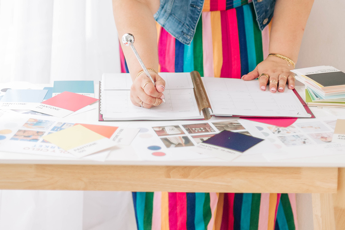 A person writes in an open planner while standing at a table covered with Pantone color cards, brand samples, and visual references. The image represents intentional planning and organization involved in building small business systems that support consistent branding and smoother workflows.