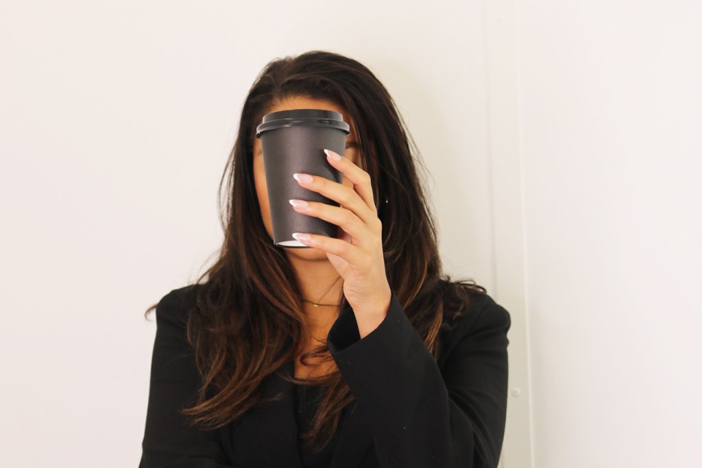 A woman in a black blazer holds a takeaway coffee cup in front of her face obscuring her expression against a neutral background. The pose suggests fatigue and reliance on caffeine often linked to entrepreneur burnout.