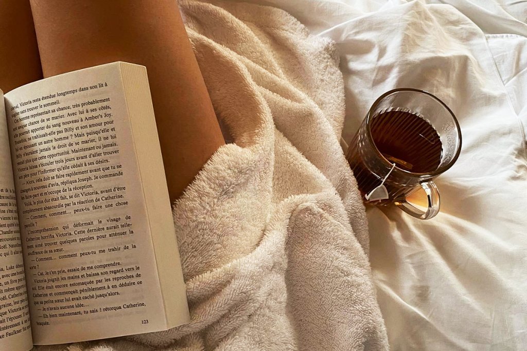 An open book with French text lies on a soft blanket beside a glass mug of tea on a bed. The cozy restful scene captures a slow intentional break from entrepreneur burnout.