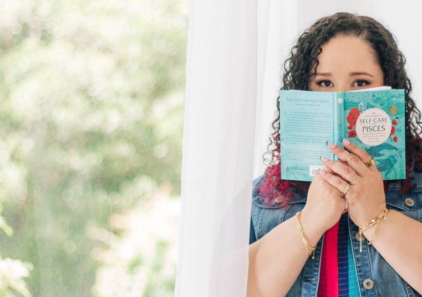 A woman stands by a bright window holding a book titled "Self-Care for Pisces" partially covering her face while looking toward the camera. The soft natural light and quiet setting reflect a personal self care moment connected to entrepreneur burnout.
