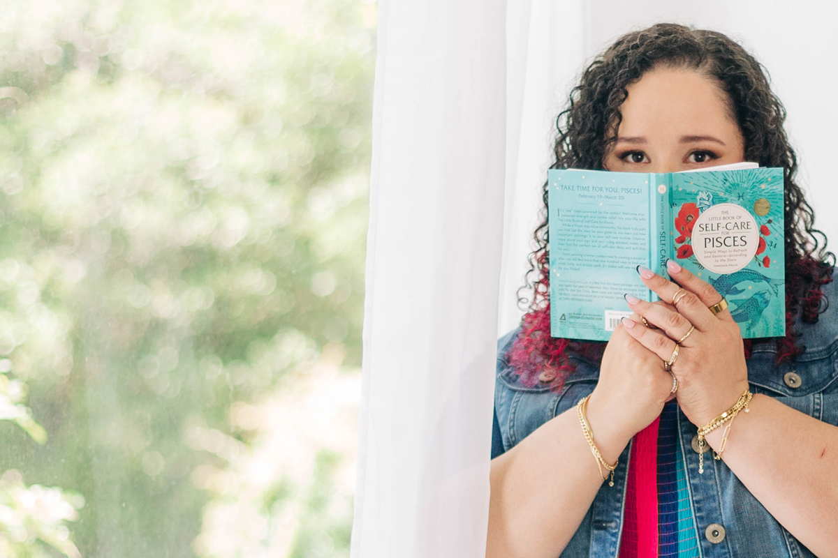 A woman stands by a bright window holding a book titled "Self-Care for Pisces" partially covering her face while looking toward the camera. The soft natural light and quiet setting reflect a personal self care moment connected to entrepreneur burnout.