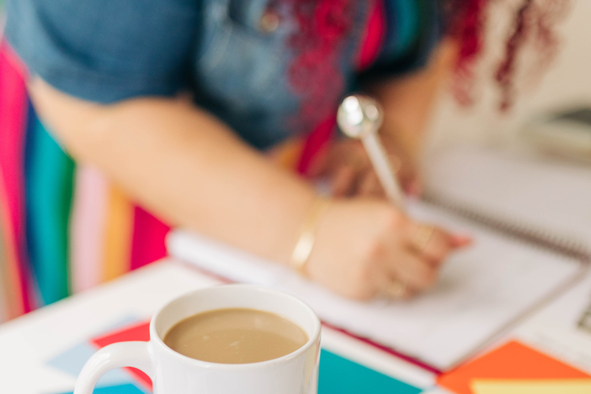 Close up of a desk with a coffee mug that reads "So, it Depends" placed on colorful branding materials labeled "SUNHOUSE SOCIAL" alongside color swatches. In the background, a person writes in a notebook while working through ideas because their website does not reflect their brand.