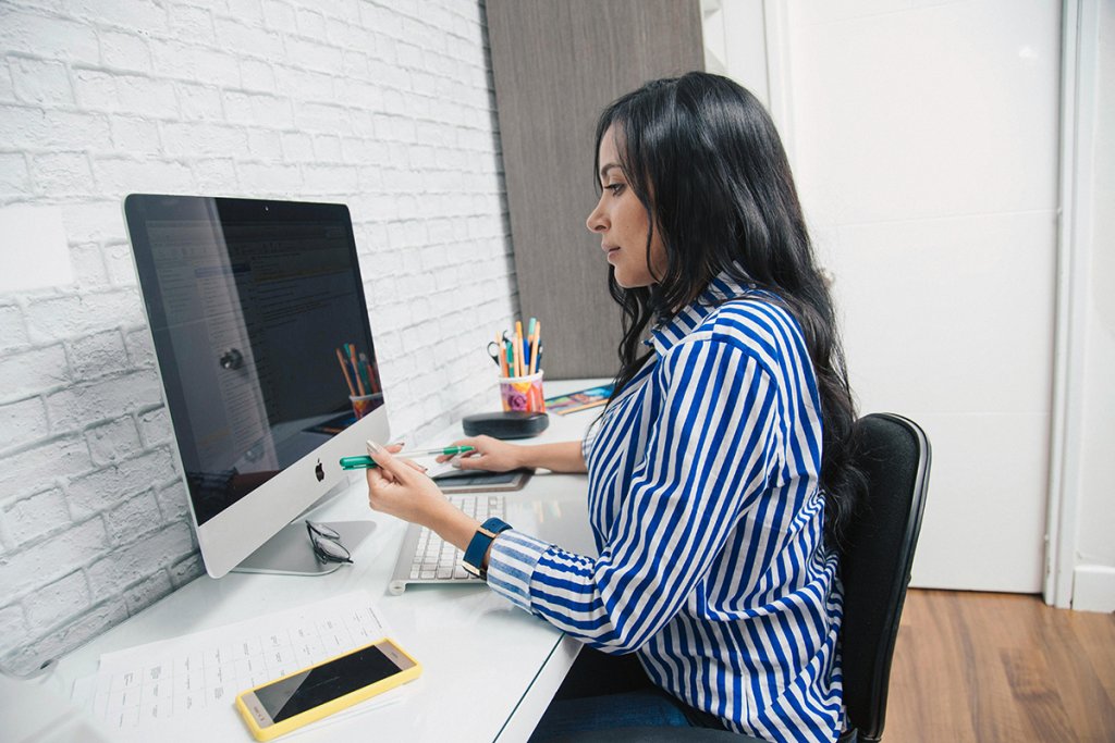 Woman in a blue striped shirt sits at a desk working on a desktop computer while holding a pen, appearing focused as she reviews content that suggests her website does not reflect her brand. A phone, glasses, and colorful pencils sit nearby in a home office setup.