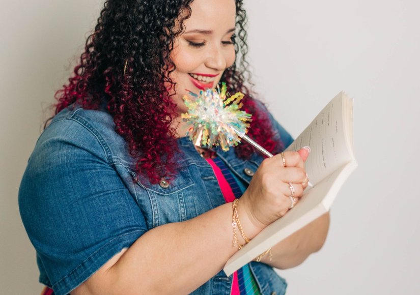 A woman in a colorful striped dress smiles while writing in a notebook with a decorative pen. The focused journaling moment reflects the process of refining website messaging and clarifying ideas.