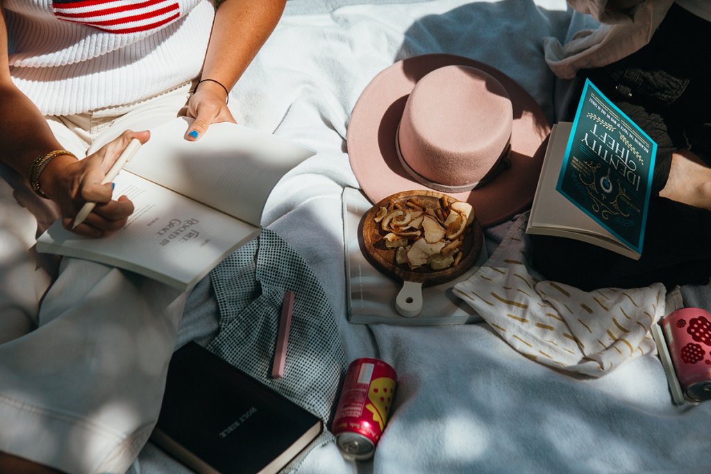 A person writes in a notebook on a bed surrounded by a hat snacks books and a drink in soft natural light. The relaxed creative setup represents brainstorming and developing website messaging.